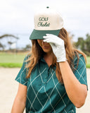 Stylish woman wearing a Golf Chalet Hat in white and green, shading her eyes with a golf glove, against a golf course backdrop.