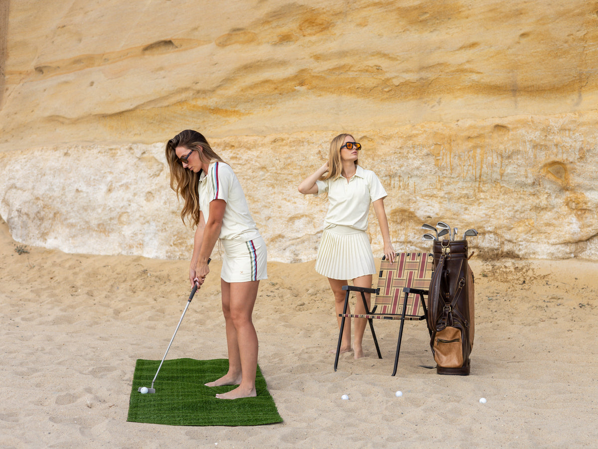 Two women on a sandy beach with a large rock face behind them, one in a white polo shirt and shorts preparing to hit a golf ball on a green turf mat, while the other stands beside a golf bag and chair.