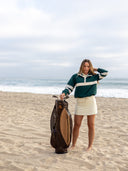 A woman stands on a sandy beach holding a golf bag, wearing a dark green and cream striped long-sleeve athletic pullover and a cream mini skirt.