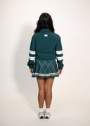 A model viewed from behind wearing a dark teal green cheerleader-style jacket and matching argyle pleated skirt with white stripes, standing against a plain off-white studio background.