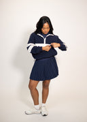 A woman modeling a navy blue and white athletic zip-up jacket and matching pleated mini skirt, paired with white sneakers, against a plain off-white studio background.