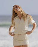 A woman wearing a cream-colored short-sleeve collared shirt and matching mini skirt stands on a beach with the ocean and sky blurred in the background.
