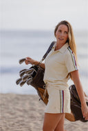 A woman on a sandy beach wearing a cream-colored polo shirt and matching shorts with thin red, green, and blue stripes down the sleeves and sides, carrying a brown golf bag over her shoulder.