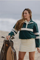 A woman on a beach wearing a dark green and cream color-blocked pullover sweater and a cream mini skirt, standing next to a brown golf bag.