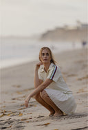 A woman crouching on a sandy beach wearing a cream-colored short-sleeve polo shirt with a thin red, blue, and green stripe detail on the sleeve, paired with a matching pleated cream skirt.