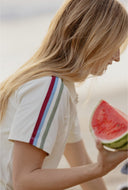 A woman in a white short-sleeve polo shirt with colorful striped details on the sleeve, holding a slice of watermelon in a beach setting.