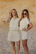 Two women standing on a sandy beach wearing matching cream-colored athletic outfits, with one in a short-sleeve polo shirt and pleated skirt and the other in a short-sleeve top and skirt with striped detailing on the sleeve.