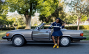 Woman in a blue dress standing next to a classic silver convertible car on a tree-lined street.
