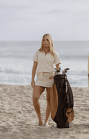 A woman stands on a sandy beach next to a dark brown and tan golf bag, wearing a cream-colored short-sleeve polo shirt and matching mini skirt.