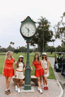 Four women in red and white outfits standing next to a large clock on a golf course.