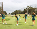 Four women in green uniforms playing golf on a course with trees and a clear sky in the background.