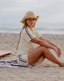 A woman sits on a striped towel on a sandy beach wearing a cream short-sleeve collared shirt and matching shorts with thin multicolor stripes, plus a wide-brim straw hat. The ocean and cloudy sky are in the background.